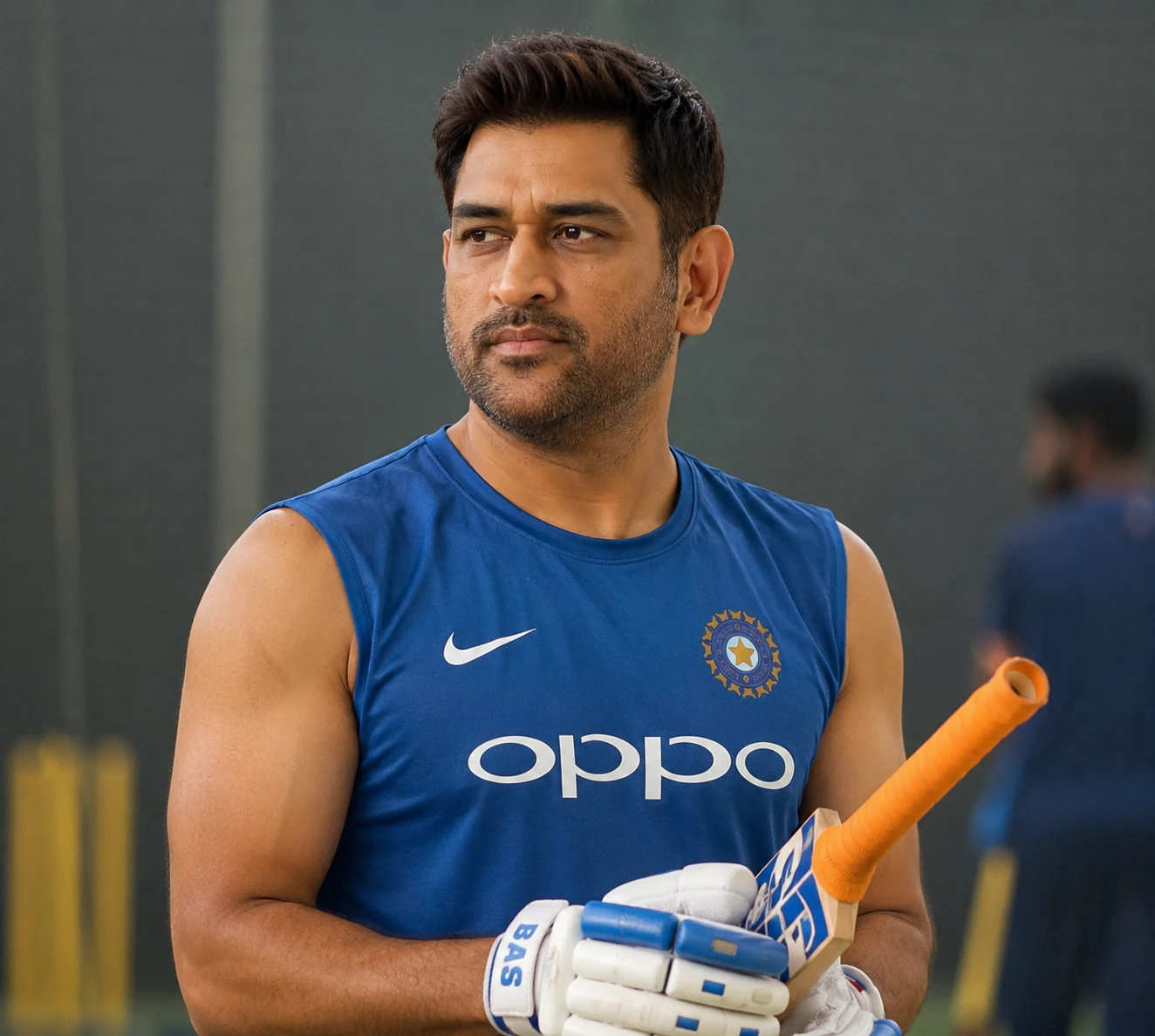 MS Dhoni practicing in indoor cricket nets wearing blue training jersey holding bat and gloves.